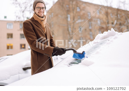 Stylish man in a coat and scarf clears snow from his car during winter snowfall. Seasonality concept Stylish man in a coat and scarf clears snow from his car during winter snowfall. Seasonality concept 109122702