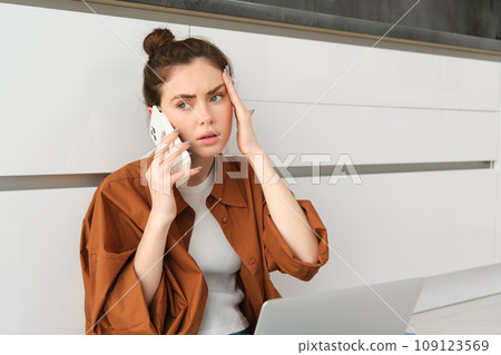 Portrait of woman calling her GP from home, holding smartphone, has headache or high fever Portrait of woman calling her GP from home, holding smartphone, has headache or high fever 109123569