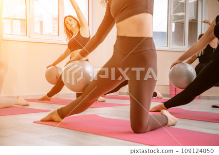 A group of six athletic women doing pilates or yoga on pink mats in front of a window in a beige loft studio interior. Teamwork, good mood and healthy lifestyle concept. A group of six athletic women doing pilates or yoga on pink mats in front of a window in a beige loft studio interior. Teamwork, good mood and healthy lifestyle concept. 109124050