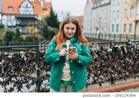 Attractive young female tourist is exploring new city. Redhead girl holding a mobile phone on city street in Gdansk. Traveling Europe in autumn. Vacation concept Attractive young female tourist is exploring new city. Redhead girl holding a mobile phone on city street in Gdansk. Traveling Europe in autumn. Vacation concept 109124364