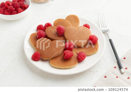A heap of heart shaped pancakes with raspberries and a fork on the white plate on white background A heap of heart shaped pancakes with raspberries and a fork on the white plate on white background 109124574
