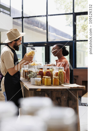 Store owner showcasing his products in marketplace venue, presenting additives free food to female customer. Local vendor selling natural organic bulk products during event to african american client. Store owner showcasing his products in marketplace venue, presenting additives free food to female customer. Local vendor selling natural organic bulk products during event to african american client. 109126332