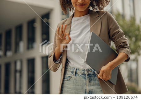 Close up of businesswoman standing with laptop on office building background and showing sign Ok Close up of businesswoman standing with laptop on office building background and showing sign Ok 109126674