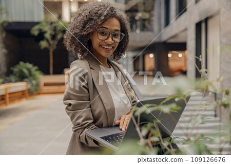 Cheerful woman freelancer working on laptop while standing in modern office and looks camera Cheerful woman freelancer working on laptop while standing in modern office and looks camera 109126676