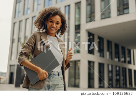 Smiling businesswoman standing with laptop on office building background and showing thumb up Smiling businesswoman standing with laptop on office building background and showing thumb up 109126683