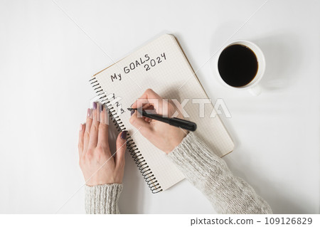 Female hands writing My Goals 2024 in a notebook. Mug of coffee on the table, top view 109126829