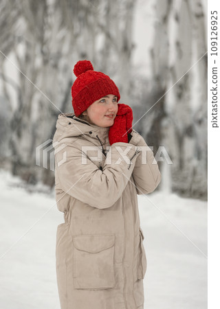Beautiful girl in warm winter clothes walks on frosty day. Young woman in down jacket and wool red hat with mittens warms up in snow park Beautiful girl in warm winter clothes walks on frosty day. Young woman in down jacket and wool red hat with mittens warms up in snow park 109126925