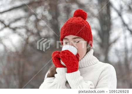 Young woman drinking hot tea or coffee from mug in nature. Brunette in beautiful knitted hat and mittens in winter park 109126928