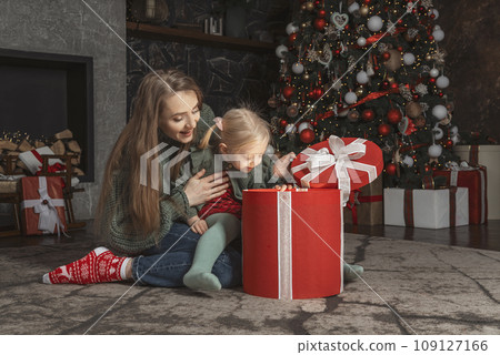 Young mother and little daughter open a gift with surprise and delight, Christmas eve. Mother and daughter near the Christmas tree. 109127166