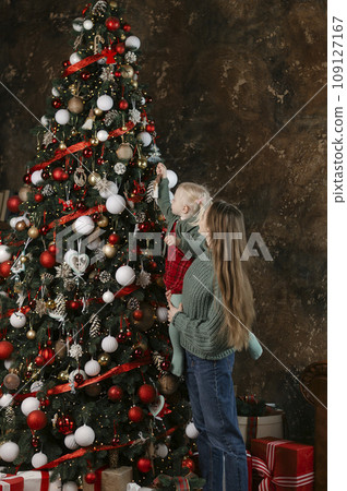 Portrait of mother with her daughter in her arms near Christmas tree. Mom and little girl decorate Christmas tree indoors. Vertical frame. Portrait of mother with her daughter in her arms near Christmas tree. Mom and little girl decorate Christmas tree indoors. Vertical frame. 109127167