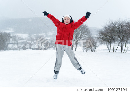 Teenage boy in red jacket and headphones jumps during snowfall. Child with his hands up and enjoys the winter holidays. Teenage boy in red jacket and headphones jumps during snowfall. Child with his hands up and enjoys the winter holidays. 109127194