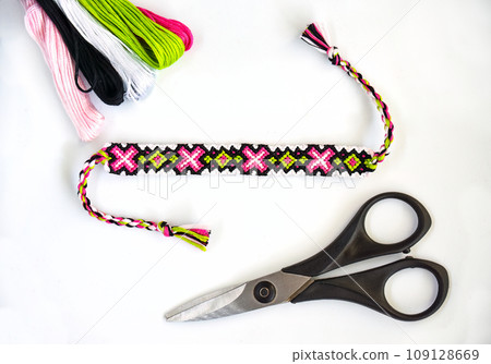 Woven handmade DIY friendship bracelets with bright pink and green pattern next to skeins of thread and scissors on white background 109128669