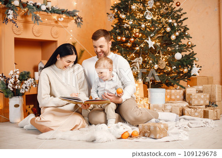 Young family celebrating Christmas together. Brunette mother, bearded father and blonde son sitting near Christmas tree and reading a book. Family wearing beige knitted clothes. 109129678
