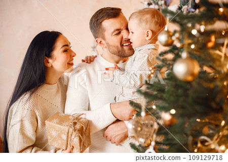 Young family celebrating Christmas together. Brunette mother, bearded father and blonde son standing near Christmas tree and holding a gift box. Family wearing beige knitted clothes. Young family celebrating Christmas together. Brunette mother, bearded father and blonde son standing near Christmas tree and holding a gift box. Family wearing beige knitted clothes. 109129718