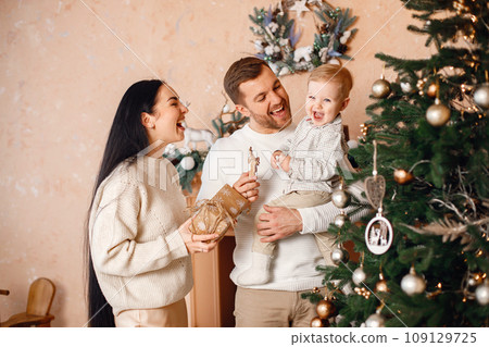 Young family celebrating Christmas together. Brunette mother, bearded father and blonde son standing near Christmas tree and holding a gift box. Family wearing beige knitted clothes. Young family celebrating Christmas together. Brunette mother, bearded father and blonde son standing near Christmas tree and holding a gift box. Family wearing beige knitted clothes. 109129725