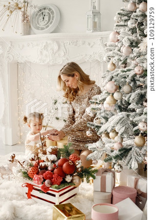 Blonde mother and little daughter in studio posing for a photo. Woman wearing golden dress and daughter white one. Family decorating Christmas together. 109129759