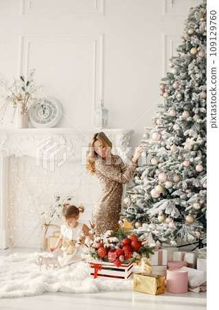 Blonde mother and little daughter in studio posing for a photo. Woman wearing golden dress and daughter white one. Family decorating Christmas together. 109129760