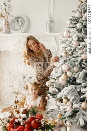 Blonde mother and little daughter in studio posing for a photo. Woman wearing golden dress and daughter white one. Family decorating Christmas together. 109129763