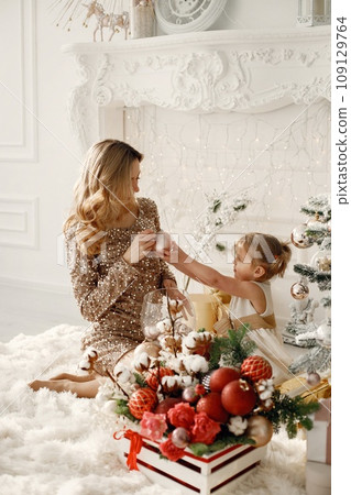 Blonde mother and little daughter in studio posing for a photo. Woman wearing golden dress and daughter white one. Family decorating Christmas together. 109129764
