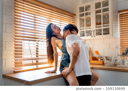 Asian man kiss on crook of the neck of woman who sit on bench in kitchen look like they enjoy sexual activity at home. 109130699