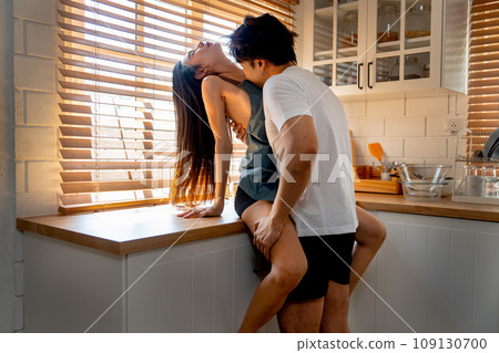 Asian man kiss on crook of the neck of woman who sit on bench in kitchen look like they enjoy sexual activity at home. 109130700