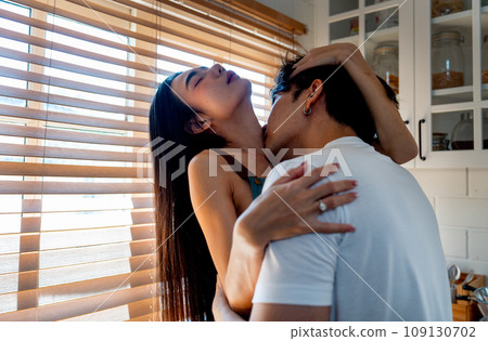 Close up of Asian man kiss on crook of the neck of woman who sit on bench in kitchen look like they enjoy sexual activity at home. 109130702