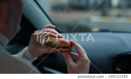 Hungry man bites piece of delicious burger satisfying hunger and sitting in vehicle in cloudy weather. Burger delivered and man eating food in vehicle . Man eating unhealthy burger in vehicle 109132395