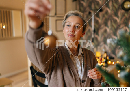 Portrait of happy middle-aged woman holding gold ball for decoration Christmas tree at home, closeup, bokeh lights, selective focus. Concept of home festive atmosphere. 109133127