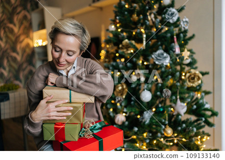 Portrait of happy elegant middle-aged woman holding big pile of wrapped gifts, sitting by decorated Christmas tree, smiling looking with happiness expression. Concept of home festive atmosphere. Portrait of happy elegant middle-aged woman holding big pile of wrapped gifts, sitting by decorated Christmas tree, smiling looking with happiness expression. Concept of home festive atmosphere. 109133140