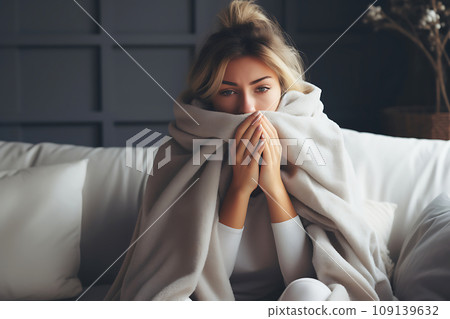 A young woman with a cold, wrapped in a blanket, sits on the sofa in the living room at home 109139632