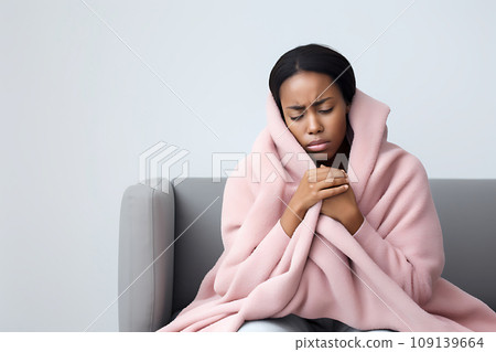 A cold and sad young African-American woman, wrapped in a pink blanket, sits on a gray sofa 109139664