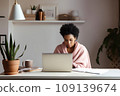 A young African-American woman, wrapped in a blanket, works remotely from home using a laptop. 109139674