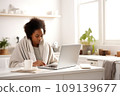 A young African-American woman, wrapped in a blanket, works remotely from home using a laptop. 109139677
