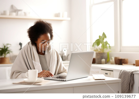 A young African-American woman, wrapped in a blanket, works remotely from home using a laptop. 109139677