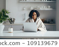 A young African-American woman, wrapped in a blanket, works remotely from home using a laptop. 109139679