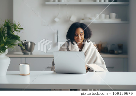 A young African-American woman, wrapped in a blanket, works remotely from home using a laptop. 109139679