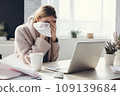 A cold young woman, wrapped in a blanket, is trying to work at home with a laptop. 109139684