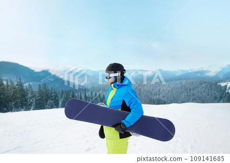 Snowboarder Of Man At Ski Resort On The Background Blue Sky,  Hold Snowboard. Wearing Ski Glasses. Ski Goggles  With The Reflection Of Snowed Mountains 109141685