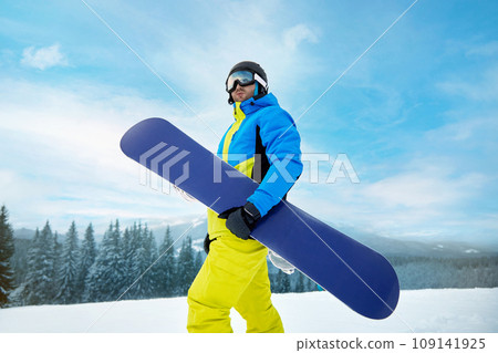 Portrait Of Man On The Background Of Mountains And Blue Sky, Holding Snowboard And Wearing Ski Glasses. Ski Goggles Of Snowboarder With The Reflection Of Snowed Mountains. Winter Sports 109141925