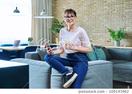 Middle aged woman with glass of water using smartphone sitting on sofa at home Middle aged woman with glass of water using smartphone sitting on sofa at home 109143962