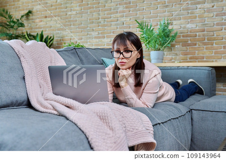 Serious middle aged woman lying at home on sofa in living room using laptop Serious middle aged woman lying at home on sofa in living room using laptop 109143964