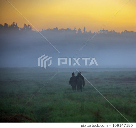 Morning autumn landscape with hunters walking in the fog through field towards the forest. Morning autumn landscape with hunters walking in the fog through field towards the forest. 109147190