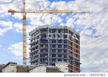 A tower crane is erecting a new residential building using the monolithic frame method against the background of the old roof of an outdated house. 109147463