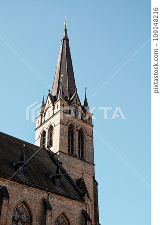 The image shows a brick church with a tall steeple and a clock on the tower. The church has arched windows and is lit by the sun against a blue sky. Spire on the bell tower of a Christian church. The image shows a brick church with a tall steeple and a clock on the tower. The church has arched windows and is lit by the sun against a blue sky. Spire on the bell tower of a Christian church. 109148216