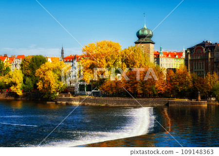 This is a photo of a river with a waterfall and buildings in the background. Ancient tower on the river bank in the fall. Autumn Prague. Landscape. This is a photo of a river with a waterfall and buildings in the background. Ancient tower on the river bank in the fall. Autumn Prague. Landscape. 109148363