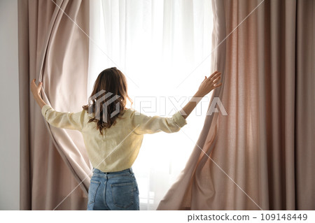Woman opening window curtains at home in morning, back view 109148449