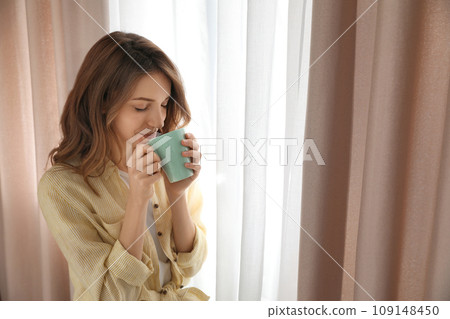 Woman drinking coffee near window with beautiful curtains at home Woman drinking coffee near window with beautiful curtains at home 109148450