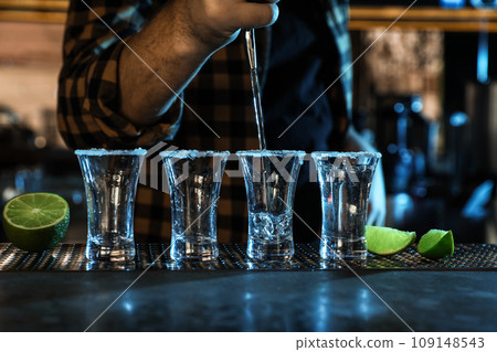 Bartender pouring Mexican Tequila into shot glasses at bar counter, closeup Bartender pouring Mexican Tequila into shot glasses at bar counter, closeup 109148543