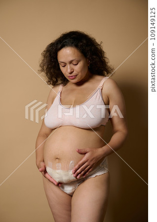 Authentic woman holding her postnatal belly with bandage hiding scar after C-section cesarean, isolated beige background Authentic woman holding her postnatal belly with bandage hiding scar after C-section cesarean, isolated beige background 109149125