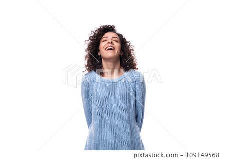authentic smiling young brunette woman with curls is dressed in a blue knitted sweater on a white 109149568